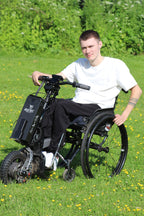 Young man using a wheelchair with Batec Mobility power attachment, wearing Vilber's adaptive joggers in black. Active lifestyle shot outdoors.