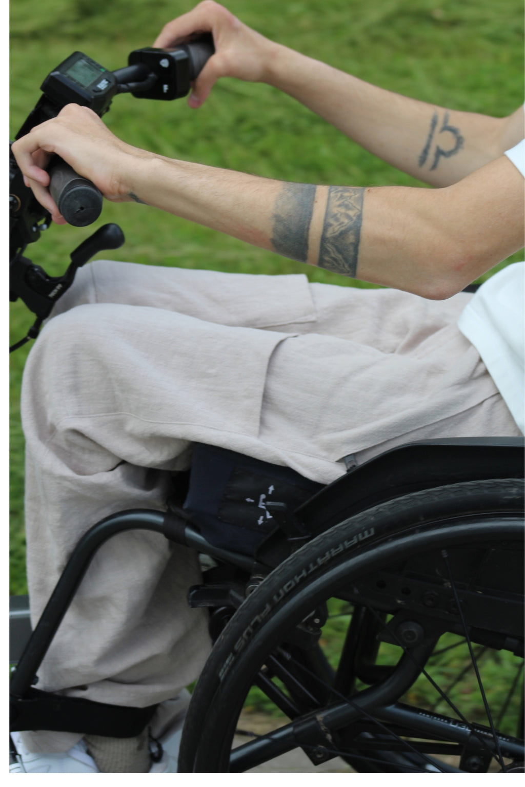 Close-up of a wheelchair user's hands on a Batec power add-on handlebar, with Vilber's beige summer trousers visible across the lap.