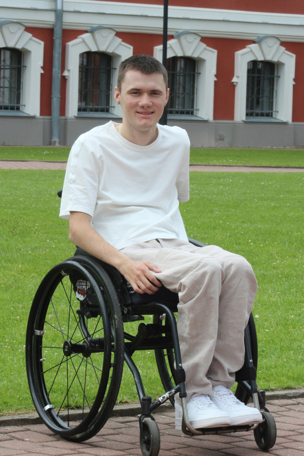 Young man sitting in a manual wheelchair outdoors, wearing Vilber's beige ramie summer trousers and a white t-shirt.